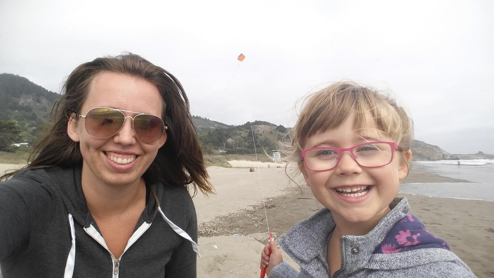 A women wearing glasses in a black hoody next to a little girl in a grey jacket holding a string connected to a flying kite behind them in the sky.