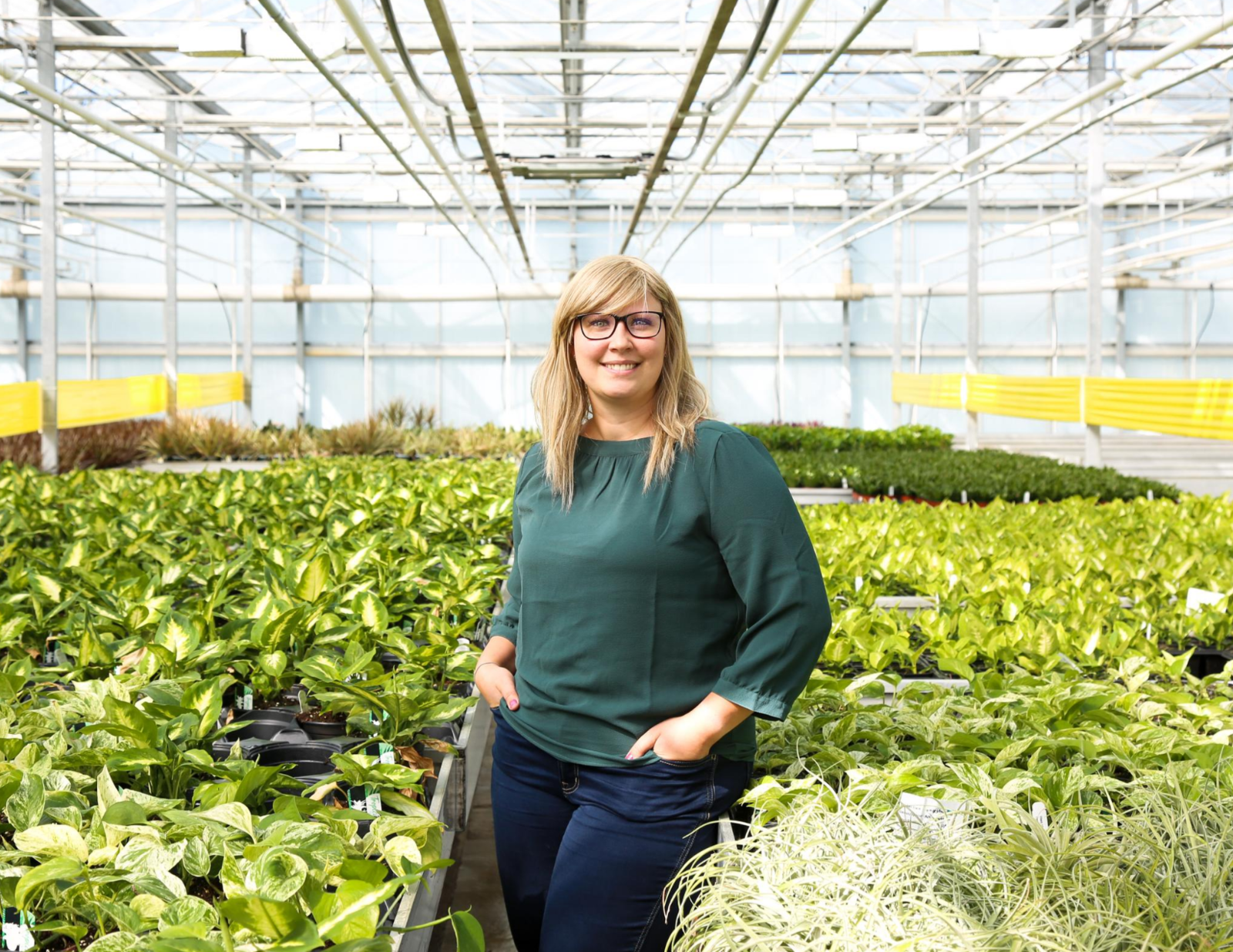 A blonde women in a turquoise shirt in the middle of a greenhouse filled with green plants.