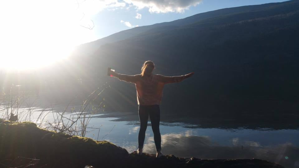 A women with hands spread out at her sides looking at a view of water and mountains below her with the sun in the left top corner shining bright.
