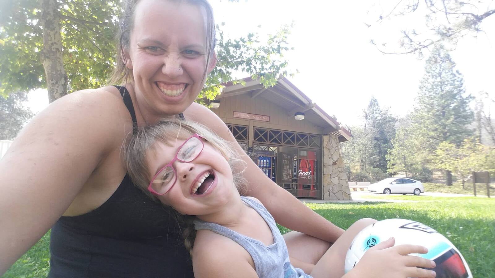 A women in a black tank holding a little girl with pink glasses who is also holding a volleyball.