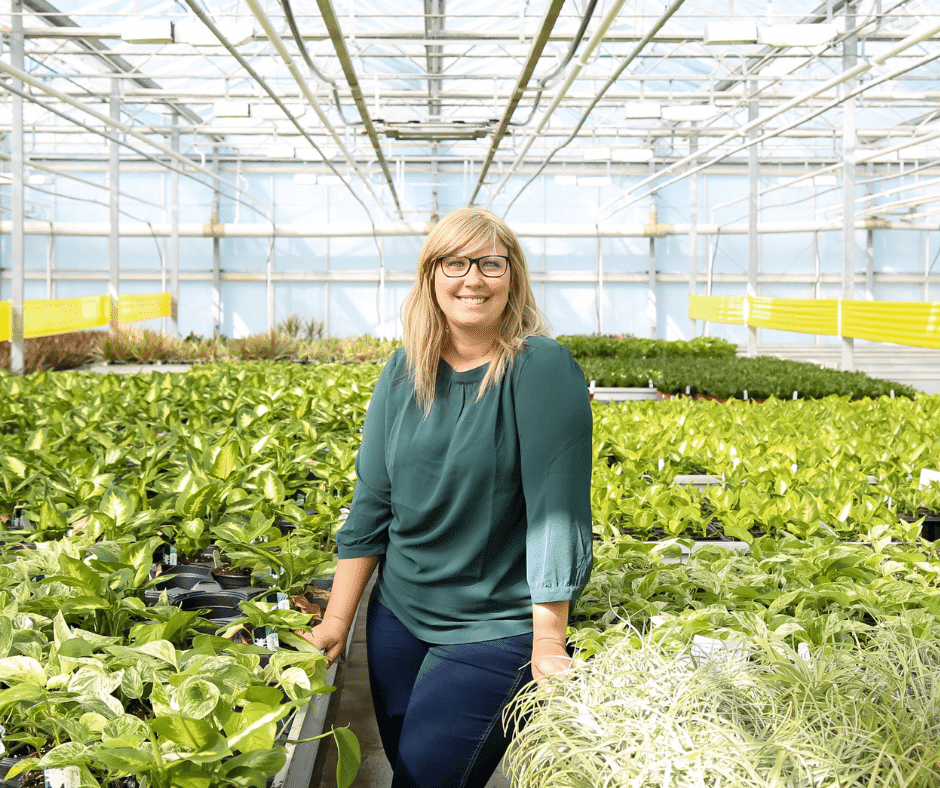 A women in a teal shirt smiling while standing in a greenhouse between plants..