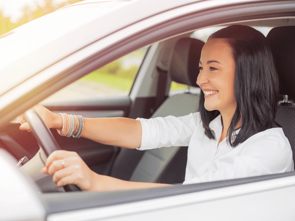 A women with brown hair wearing a white shirt in a white car smiling as she drives