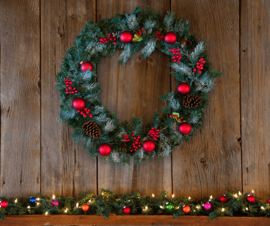  A Green wreath hung up on a wooden wall with red bulbs and red berries and brown pinecones on it, with multicolored light garland down below.