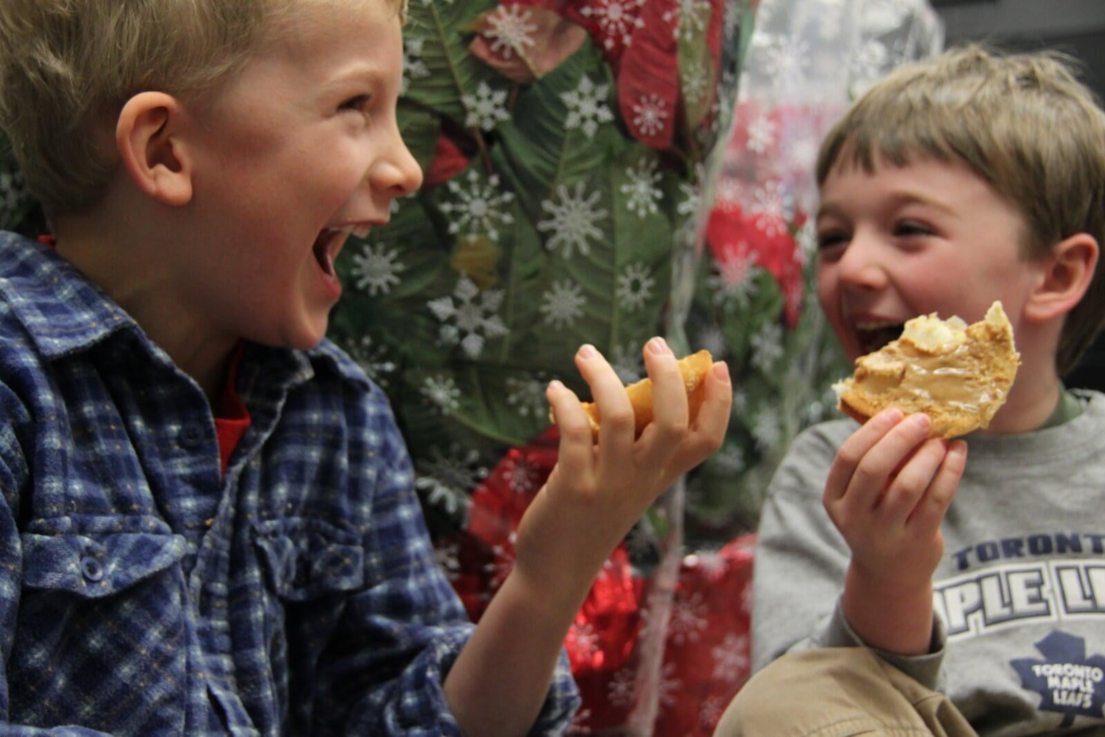 Two boys sitting laughing eating bagels in front of red packaged poinsettias.